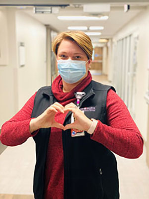 a nurse wearing a fask mask while standing in a hospital hallway holds their hands in the shape of a heart